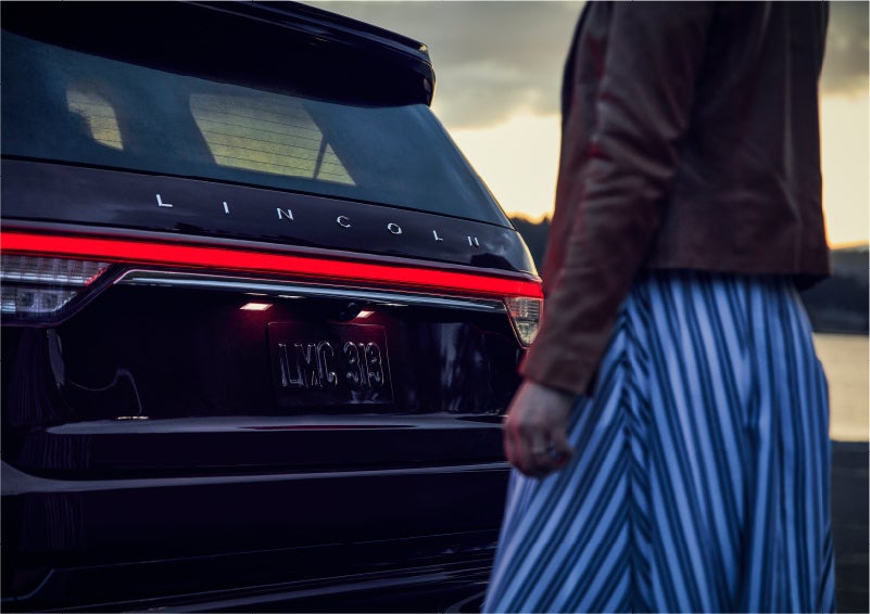 A person is shown near the rear of a 2023 Lincoln Aviator® SUV as the Lincoln Embrace illuminates the rear lights | Bozard Lincoln in Saint Augustine FL