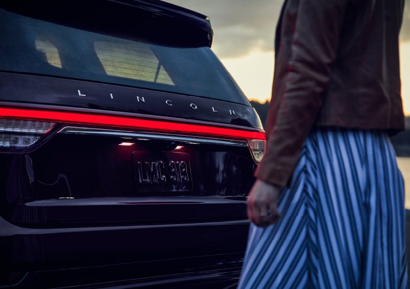 A person is shown near the rear of a 2024 Lincoln Aviator® SUV as the Lincoln Embrace illuminates the rear lights | Bozard Lincoln in Saint Augustine FL